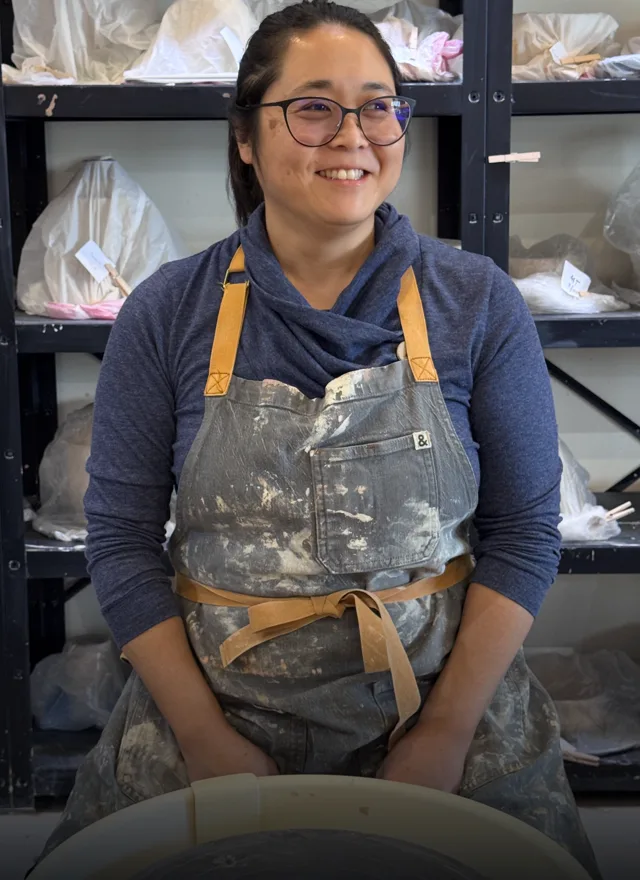Dr. Jennifer Fan sitting in front of a potter's wheel wearing an apron.
